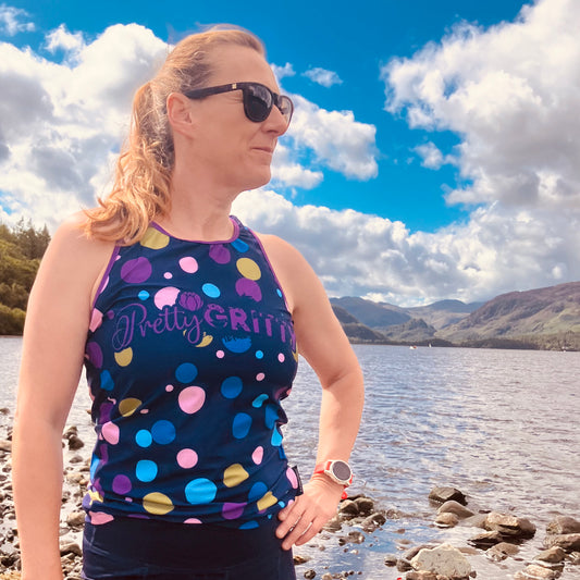 A woman wearing a colorful polka-dot tank top with 'Pretty GRITTY' printed on the front, standing on a rocky lakeshore with mountains and a cloudy blue sky in the background.
